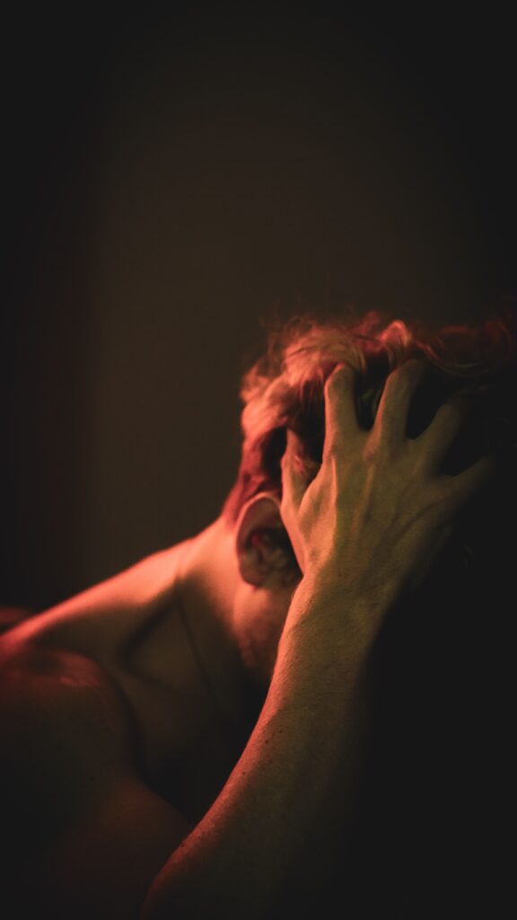 A shirtless man in dramatic lighting with hands in hair, evoking a sense of introspection.
