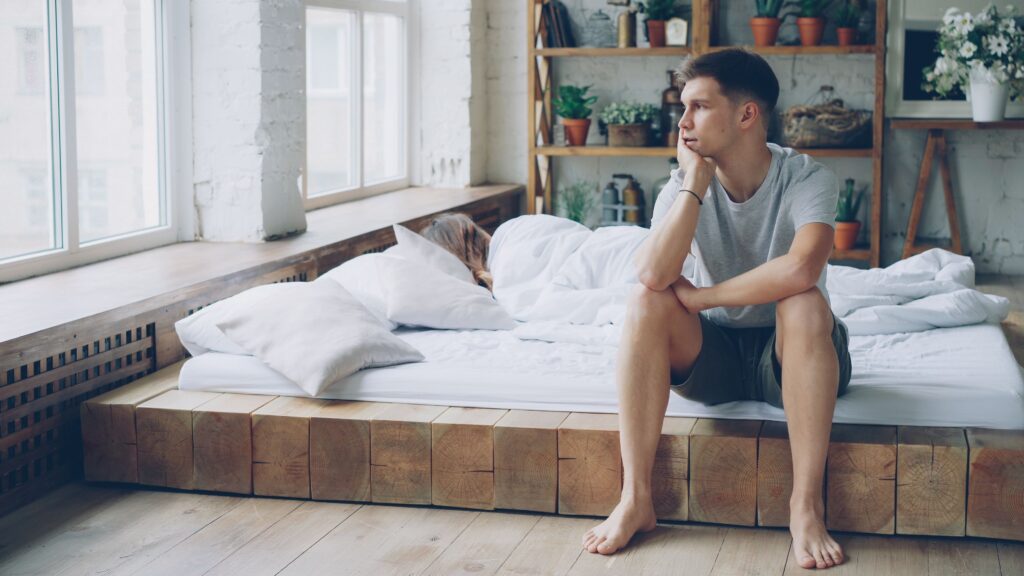 A thoughtful young man sits by a window in a cozy, modern bedroom.