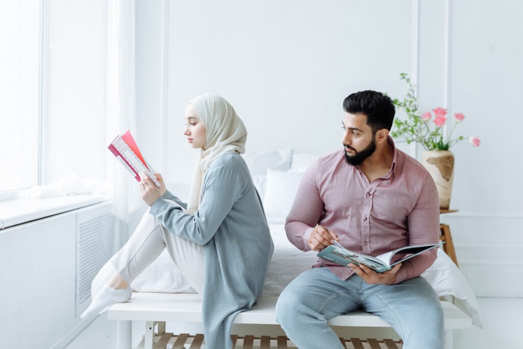 A couple seated on a bed, reading books in a bright, cozy room.